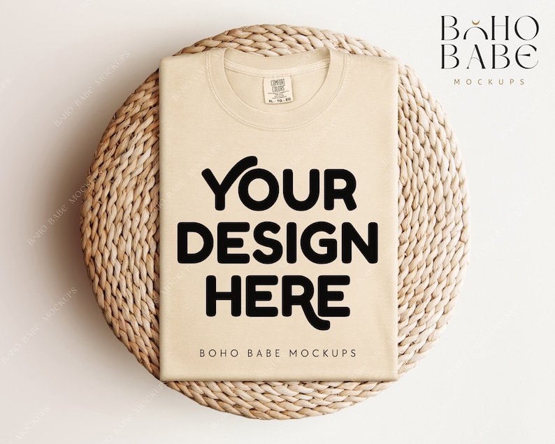 May include: A beige t-shirt mockup on a woven straw mat. The t-shirt has the text "YOUR DESIGN HERE" printed on the front. The text "BOHO BABE MOCKUPS" is printed below the design.
