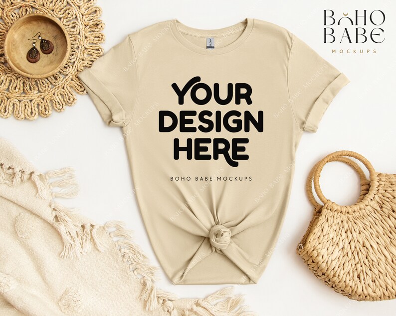 May include: A beige t-shirt with short sleeves and a crew neck. The shirt is knotted at the bottom and has the text "YOUR DESIGN HERE" printed on the front. The shirt is laid flat on a white surface with a woven basket and a blanket.