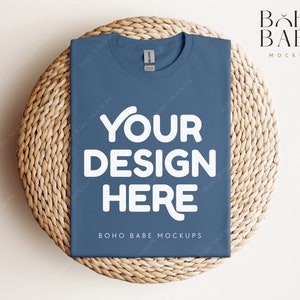 Op de afbeelding: Een blauwe t-shirt mockup op een geweven strooien mat. Het t-shirt heeft de tekst "YOUR DESIGN HERE" in wit. De tekst "BOHO BABE MOCKUPS" is ook zichtbaar op het t-shirt.