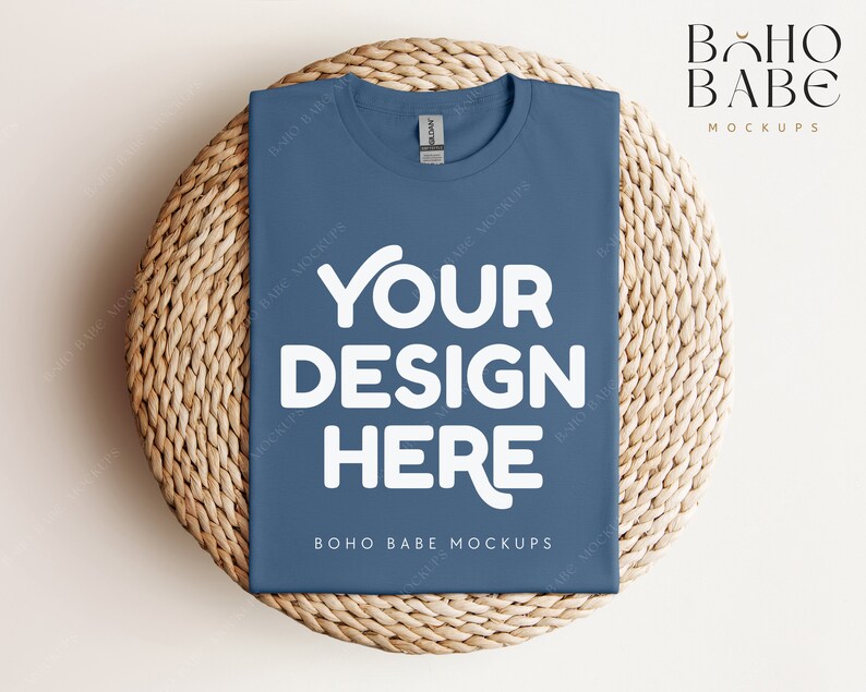Op de afbeelding: Een blauwe t-shirt mockup op een geweven strooien mat. Het t-shirt heeft de tekst "YOUR DESIGN HERE" in wit. De tekst "BOHO BABE MOCKUPS" is ook zichtbaar op het t-shirt.