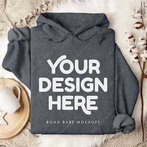 Puede incluir: Una sudadera con capucha de color gris oscuro con el texto "YOUR DESIGN HERE" y "BOHO BABE MOCKUPS" impreso en la parte delantera. La sudadera est&aacute; colocada sobre una superficie de piel sint&eacute;tica blanca.