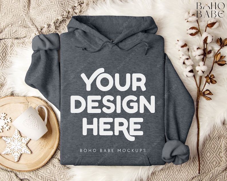 Puede incluir: Una sudadera con capucha de color gris oscuro con el texto "YOUR DESIGN HERE" y "BOHO BABE MOCKUPS" impreso en la parte delantera. La sudadera est&aacute; colocada sobre una superficie de piel sint&eacute;tica blanca.