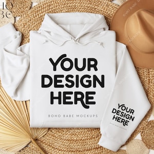 Op de afbeelding: Een witte hoodie met de tekst "YOUR DESIGN HERE" in vette zwarte letters op de voorkant en mouw. De hoodie wordt gepresenteerd op een geweven mat met een bruine hoed en decoratieve elementen.