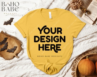 May include: A yellow t-shirt with the text "YOUR DESIGN HERE" in black. The shirt is laid out on a white knitted blanket with autumnal decor, including a basket of pumpkins and a book.