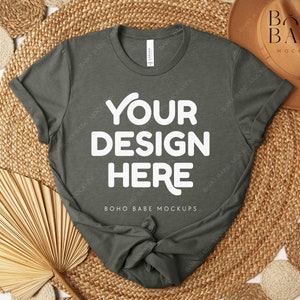 Peut inclure: Un t-shirt vert fonc&eacute; avec le texte "YOUR DESIGN HERE" en grandes lettres blanches. Le t-shirt est pr&eacute;sent&eacute; sur un tapis tiss&eacute; avec des &eacute;l&eacute;ments d&eacute;coratifs, dont un chapeau et des feuilles de palmier s&eacute;ch&eacute;es. Les mots "BOHO BABE MOCKUPS" sont &eacute;galement visibles.