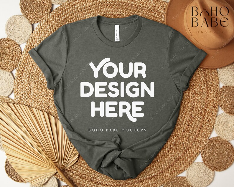 Peut inclure: Un t-shirt vert fonc&eacute; avec le texte "YOUR DESIGN HERE" en grandes lettres blanches. Le t-shirt est pr&eacute;sent&eacute; sur un tapis tiss&eacute; avec des &eacute;l&eacute;ments d&eacute;coratifs, dont un chapeau et des feuilles de palmier s&eacute;ch&eacute;es. Les mots "BOHO BABE MOCKUPS" sont &eacute;galement visibles.