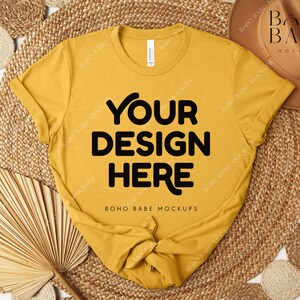 Peut inclure: T-shirt jaune moutarde avec le texte "YOUR DESIGN HERE" en lettres noires grasses. Le t-shirt est nou&eacute; en bas. L'image comprend un chapeau marron, des dessous de plat tiss&eacute;s et une feuille de palmier s&eacute;ch&eacute;e.
