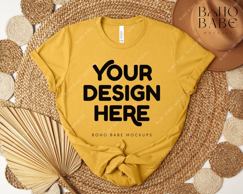 Peut inclure: T-shirt jaune moutarde avec le texte "YOUR DESIGN HERE" en lettres noires grasses. Le t-shirt est nou&eacute; en bas. L'image comprend un chapeau marron, des dessous de plat tiss&eacute;s et une feuille de palmier s&eacute;ch&eacute;e.