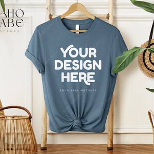 Peut inclure: Une maquette de t-shirt bleu avec le texte "Your Design Here" et "Boho Babe Mockups" sur un fond blanc. Le t-shirt est suspendu à un cintre en bois.