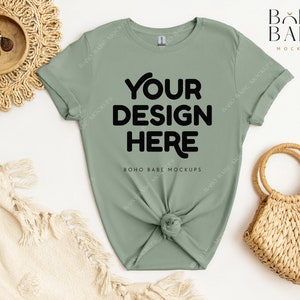 May include: A sage green t-shirt with short sleeves and a knotted front. The shirt has the text "YOUR DESIGN HERE" printed in black on the front. The shirt is laid flat on a white surface with a woven basket and a blanket.