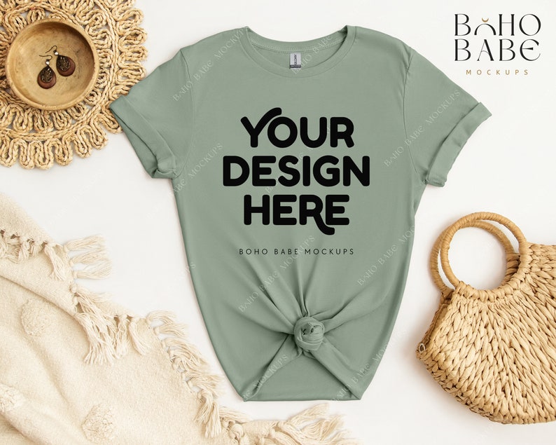 May include: A sage green t-shirt with short sleeves and a knotted front. The shirt has the text "YOUR DESIGN HERE" printed in black on the front. The shirt is laid flat on a white surface with a woven basket and a blanket.