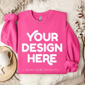 以下が含まれることがあります： ピンクのトレーナーに、白い文字で「YOUR DESIGN HERE」と書いてあります。トレーナーは、白いふわふわした表面に置かれており、左側に木のトレイと白いマグカップがあります。