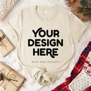 May include: A cream-colored t-shirt with the text "YOUR DESIGN HERE" in bold black letters. The shirt is laid on a white fluffy surface, with wrapped gifts and holiday-themed accessories. The shirt is a mock-up.