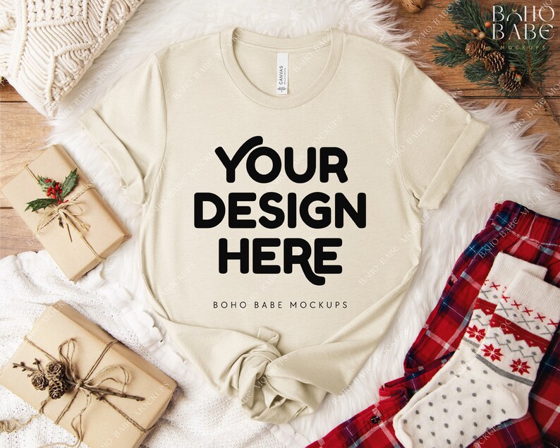 May include: A cream-colored t-shirt with the text "YOUR DESIGN HERE" in bold black letters. The shirt is laid on a white fluffy surface, with wrapped gifts and holiday-themed accessories. The shirt is a mock-up.
