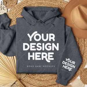 Op de afbeelding: Een donkergrijze hoodie met de tekst "YOUR DESIGN HERE" op de voorkant en "YOUR DESIGN HERE" op de mouw. De hoodie ligt op een geweven strooien mat met een strooien waaier en een bruine hoed.