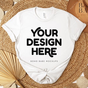 Peut inclure: Un t-shirt blanc avec le texte "YOUR DESIGN HERE" en lettres noires. Le t-shirt est pr&eacute;sent&eacute; sur un tapis tiss&eacute;, avec un chapeau marron et d'autres &eacute;l&eacute;ments d&eacute;coratifs. Les mots "BOHO BABE MOCKUPS" sont &eacute;galement visibles.