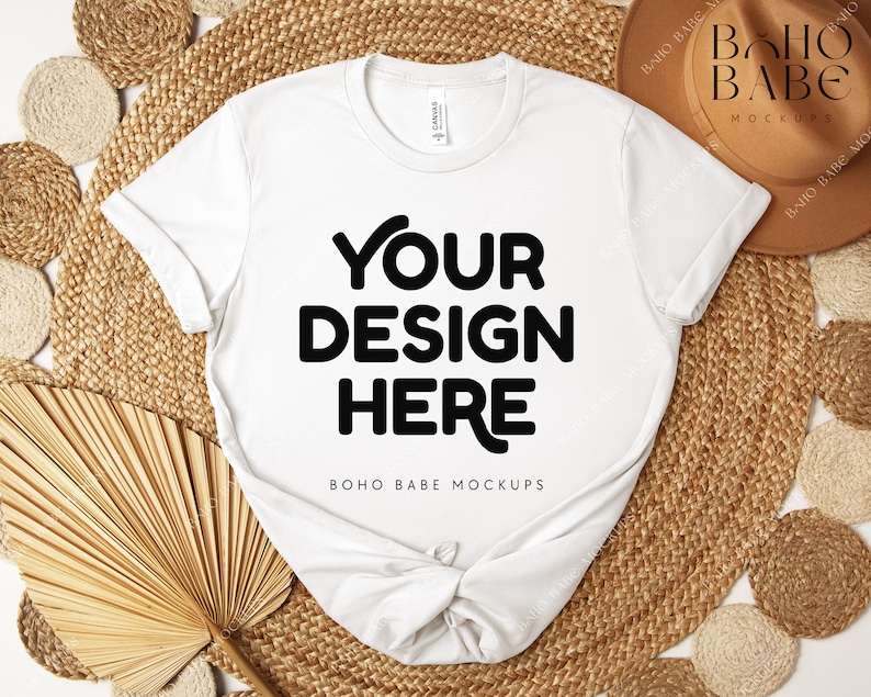 Peut inclure: Un t-shirt blanc avec le texte "YOUR DESIGN HERE" en lettres noires. Le t-shirt est pr&eacute;sent&eacute; sur un tapis tiss&eacute;, avec un chapeau marron et d'autres &eacute;l&eacute;ments d&eacute;coratifs. Les mots "BOHO BABE MOCKUPS" sont &eacute;galement visibles.