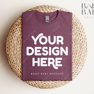 Op de afbeelding: Een mock-up van een maroon t-shirt met de tekst "YOUR DESIGN HERE" in wit op een geweven strooien onderzetter. De tekst "BOHO BABE MOCKUPS" is ook zichtbaar op het t-shirt.