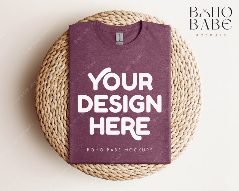 Op de afbeelding: Een mock-up van een maroon t-shirt met de tekst "YOUR DESIGN HERE" in wit op een geweven strooien onderzetter. De tekst "BOHO BABE MOCKUPS" is ook zichtbaar op het t-shirt.