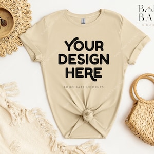 May include: A beige t-shirt with short sleeves and a crew neck. The shirt is knotted at the bottom and has the text "YOUR DESIGN HERE" printed on the front. The shirt is laid flat on a white surface with a woven basket and a blanket.
