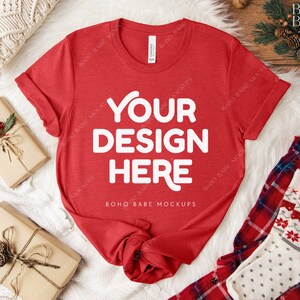 May include: A red t-shirt with the text "YOUR DESIGN HERE" in white, displayed on a white fur-like surface. The shirt is accompanied by wrapped gifts, a pair of plaid pajama pants, and a pair of patterned socks. The scene is set on a wooden surface.