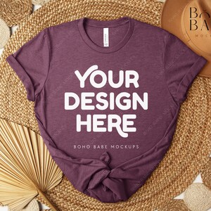 Peut inclure: Un t-shirt bordeaux avec le texte "YOUR DESIGN HERE" en lettres blanches. Le t-shirt est pr&eacute;sent&eacute; sur un tapis tiss&eacute; avec des &eacute;l&eacute;ments d&eacute;coratifs, dont un chapeau et des feuilles de palmier s&eacute;ch&eacute;es. Les mots "BOHO BABE MOCKUPS" sont &eacute;galement visibles.