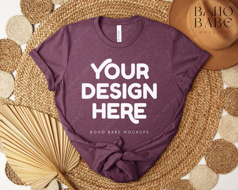 Peut inclure: Un t-shirt bordeaux avec le texte "YOUR DESIGN HERE" en lettres blanches. Le t-shirt est pr&eacute;sent&eacute; sur un tapis tiss&eacute; avec des &eacute;l&eacute;ments d&eacute;coratifs, dont un chapeau et des feuilles de palmier s&eacute;ch&eacute;es. Les mots "BOHO BABE MOCKUPS" sont &eacute;galement visibles.
