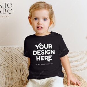 以下が含まれることがあります： 白い文字で「YOUR DESIGN HERE」と書かれた黒いTシャツを着た幼い子供。 このシャツはデザインを紹介するのに最適なモックアップです。 子供は織りのクッションに座っており、背景にはマクラメの壁掛けがあります。