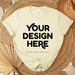 Peut inclure: Un t-shirt cr&egrave;me avec le texte "YOUR DESIGN HERE" en lettres noires en gras. Le t-shirt est pr&eacute;sent&eacute; sur un tapis tiss&eacute; avec un chapeau marron et des &eacute;l&eacute;ments d&eacute;coratifs. Les mots "BOHO BABE MOCKUPS" sont &eacute;galement visibles.