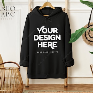May include: A black hoodie with the text "YOUR DESIGN HERE" in white. The hoodie is hanging on a wooden hanger against a white wall. A woven basket and a round straw bag are also visible.