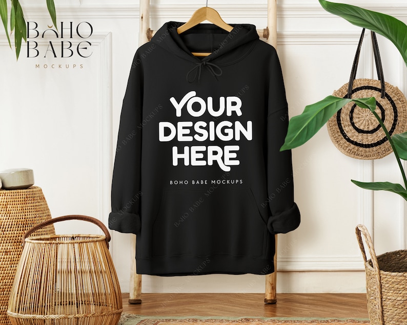 May include: A black hoodie with the text "YOUR DESIGN HERE" in white. The hoodie is hanging on a wooden hanger against a white wall. A woven basket and a round straw bag are also visible.