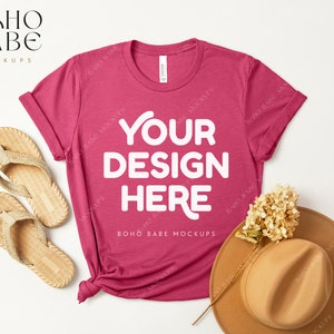 Puede incluir: Una camiseta rosa con el texto "YOUR DESIGN HERE" en blanco. La camiseta está colocada sobre una superficie blanca con un sombrero de paja y sandalias.