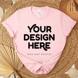 Peut inclure: Un t-shirt rose clair avec l'inscription "YOUR DESIGN HERE" en grosses lettres noires. Le t-shirt est pr&eacute;sent&eacute; sur un tapis tiss&eacute;, avec un chapeau marron et d'autres &eacute;l&eacute;ments d&eacute;coratifs. Le texte "BOHO BABE MOCKUPS" est &eacute;galement visible.