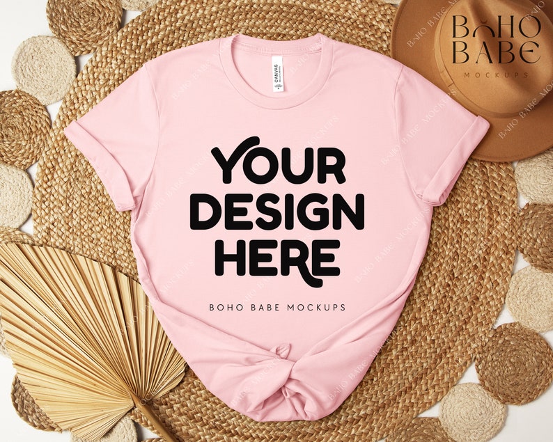 Peut inclure: Un t-shirt rose clair avec l'inscription "YOUR DESIGN HERE" en grosses lettres noires. Le t-shirt est pr&eacute;sent&eacute; sur un tapis tiss&eacute;, avec un chapeau marron et d'autres &eacute;l&eacute;ments d&eacute;coratifs. Le texte "BOHO BABE MOCKUPS" est &eacute;galement visible.