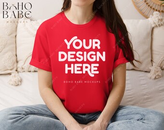 May include: A red t-shirt with the text "YOUR DESIGN HERE" in white. The shirt is being worn by a person sitting on a bed with white bedding.