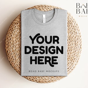 Może przedstawiać: Makieta szarego t-shirtu z nadrukiem "YOUR DESIGN HERE" na tle z plecionego wikliny.  BOHO BABE MOCKUPS jest nadrukowane na koszulce.