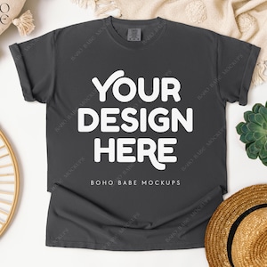 May include: A dark gray t-shirt with the text "YOUR DESIGN HERE" in white. The shirt is laid flat on a white surface, with a straw hat, a succulent plant, and a decorative woven item nearby. The words "BOHO BABE MOCKUPS" are also visible.