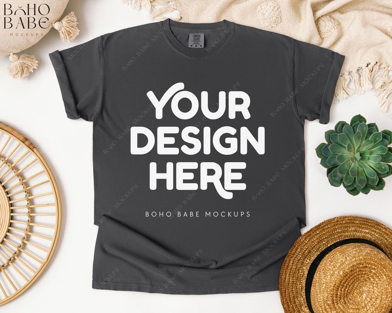 May include: A dark gray t-shirt with the text "YOUR DESIGN HERE" in white. The shirt is laid flat on a white surface, with a straw hat, a succulent plant, and a decorative woven item nearby. The words "BOHO BABE MOCKUPS" are also visible.