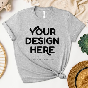 May include: Gray t-shirt mockup with the text "YOUR DESIGN HERE" and "BOHO BABE MOCKUPS" on a white background with a straw hat, a succulent plant, and a woven basket.
