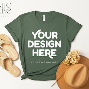 May include: A green t-shirt with the text "YOUR DESIGN HERE" in white. The t-shirt is laid flat on a white surface with a straw hat, a sprig of dried flowers, and two pairs of sandals.