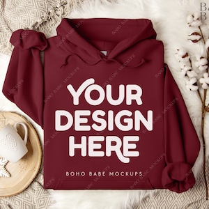 May include: A maroon hoodie with the text "YOUR DESIGN HERE" in white. The hoodie is laid on a white fur-like surface, with a wooden coaster, a white mug, and decorative snowflakes to the left. Cotton stems are on the right.