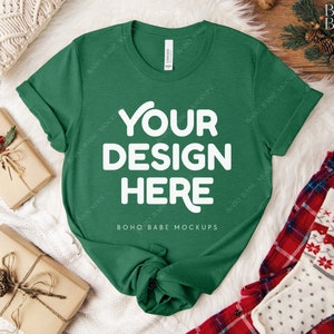May include: A green t-shirt with the text "YOUR DESIGN HERE" in white, displayed on a white fur-like surface. The shirt is surrounded by wrapped gifts, a pair of red plaid pajama pants, and a pair of Christmas socks.
