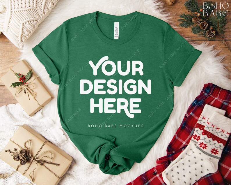 May include: A green t-shirt with the text "YOUR DESIGN HERE" in white, displayed on a white fur-like surface. The shirt is surrounded by wrapped gifts, a pair of red plaid pajama pants, and a pair of Christmas socks.