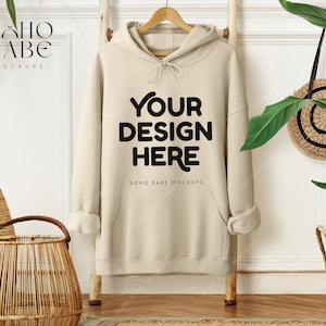 May include: A beige hoodie with the text "YOUR DESIGN HERE" in black, hanging on a wooden ladder. The hoodie is part of a mockup, with the brand name "BOHO BABE MOCKUPS" visible. The scene includes decorative baskets and plants.