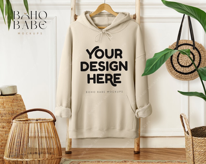 May include: A beige hoodie with the text "YOUR DESIGN HERE" in black, hanging on a wooden ladder. The hoodie is part of a mockup, with the brand name "BOHO BABE MOCKUPS" visible. The scene includes decorative baskets and plants.