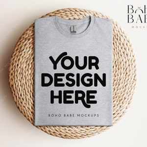 Op de afbeelding: Een mockup van een grijs t-shirt op een geweven strooien onderzetter. De tekst "YOUR DESIGN HERE" is in zwart op het shirt gedrukt. De tekst "BOHO BABE MOCKUPS" is in zwart onder het ontwerp gedrukt.