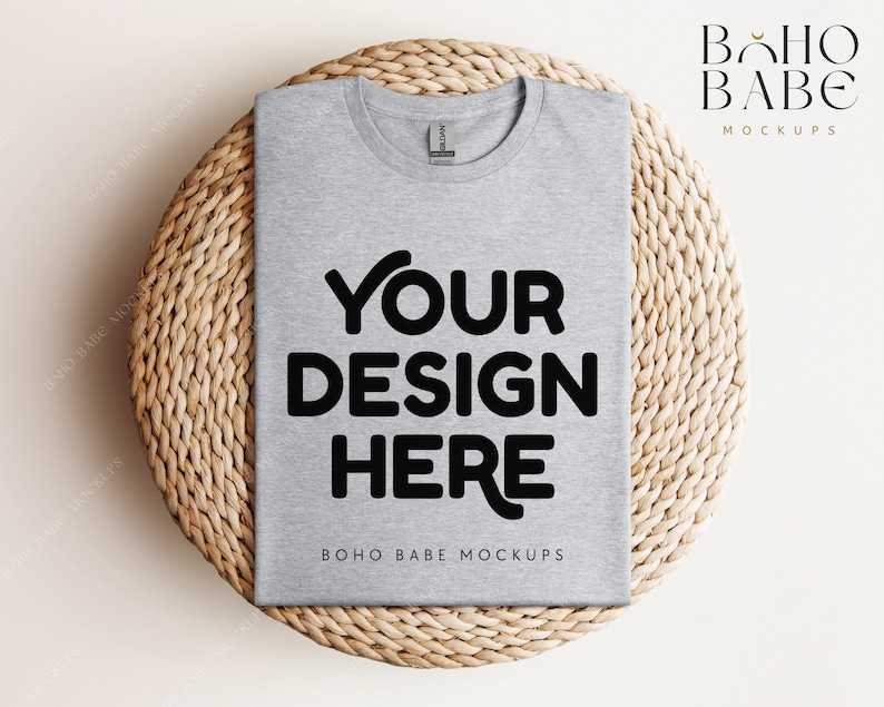 Op de afbeelding: Een mockup van een grijs t-shirt op een geweven strooien onderzetter. De tekst "YOUR DESIGN HERE" is in zwart op het shirt gedrukt. De tekst "BOHO BABE MOCKUPS" is in zwart onder het ontwerp gedrukt.