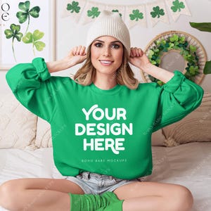 May include: A green sweatshirt with the text "YOUR DESIGN HERE" and "BOHO BABE MOCKUPS" printed on the front. The sweatshirt is being modelled by a person wearing a white beanie and sitting on a bed with a white blanket.