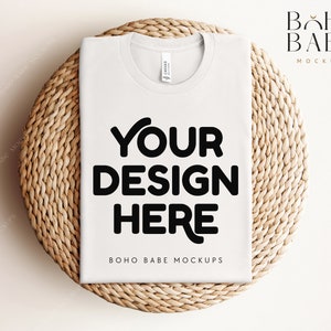 Puede incluir: Maqueta de camiseta blanca sobre una estera redonda tejida. La camiseta tiene el texto "YOUR DESIGN HERE" y "BOHO BABE MOCKUPS" impreso en ella.