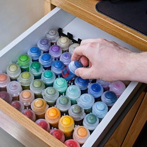 Puede incluir: Un cajón lleno de pequeños botes de pintura de varios colores, incluyendo rojo, naranja, amarillo, verde, azul y morado. Una mano alcanza un bote azul y blanco. El cajón está dentro de un mueble de madera.